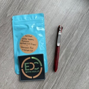 Blue zip pouch with a tan circular label, an 'EDC with Sully' card, and a red-handled utility knife on a wooden surface.