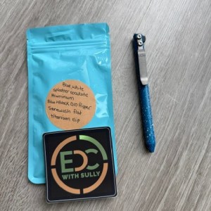 Blue resealable bag with a beige circular label, a square patch reading 'EDC WITH SULLY', and a blue glittery pen on a light gray wooden surface.