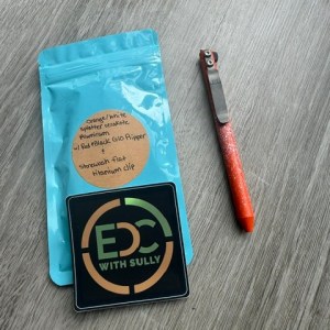 Blue resealable bag with an orange circular label and a black 'EDC with Sully' sticker, beside an orange utility knife on a gray surface.
