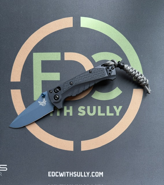 Blue folding knife with a gray textured handle and black screws laid over an orange-green circular 'EDC with Sully' logo.