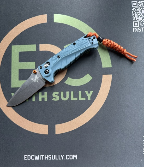 Blue folding knife with orange lanyard resting on a black surface featuring the 'EDC with Sully' logo and circle graphic.