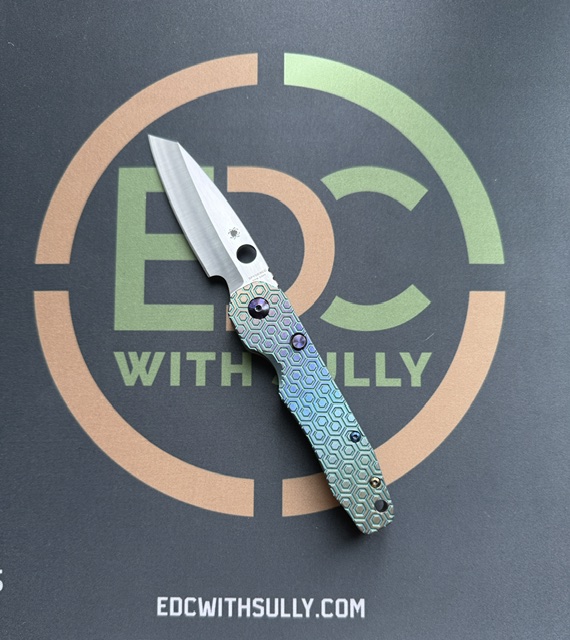 Folding knife with a honeycomb-patterned blue handle resting on a dark surface with the EDC With Sully logo.